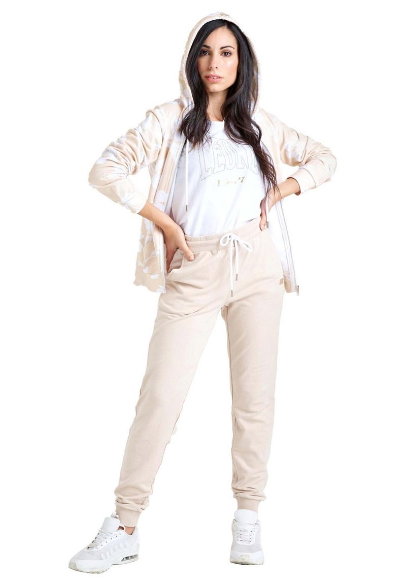 Une femme porte une veste à capuche beige clair et un pantalon de jogging avec des baskets blanches sur un fond blanc uni.