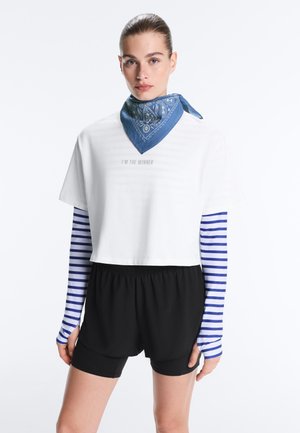 Vrouw draagt een witte cropped T-shirt over een blauw-wit gestreept shirt met lange mouwen, zwarte short en een blauw sjaaltje met patroon om de hals.