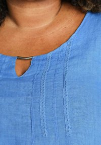 Gros plan d'une personne portant un chemisier bleu texturé avec une bordure verticale en dentelle et un petit décolleté en forme de trou de serrure maintenu par une fine barre métallique.