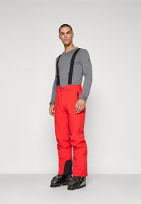 Pantalones de esquí rojos con tirantes negros ajustables, un bolsillo con cremallera, puños negros reforzados, combinados con botas de esquí negras y una camiseta de manga larga gris.