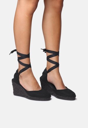 Juodos wedge espadrilės su tekstūruota pintine padu. Yra kryžminiai kojų raiščiai ir lygus kanvas viršus. Atvira kulno konstrukcija.