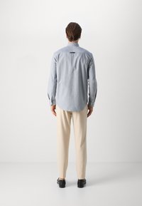 Muž stojící zády k fotoaparátu, oblečený v světle modré košili s dlouhým rukávem, béžových kalhotách, černých botách a stříbrných kruhových náušnicích, na jednobarevném pozadí.