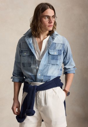 Jeune personne aux longs cheveux portant des lunettes rondes, une chemise en denim patchwork, un short blanc, un pull bleu marine noué à la taille et une montre-bracelet.