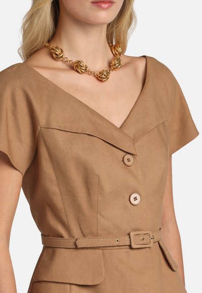 Woman wearing a brown belted dress with short sleeves and large buttons, accessorized with a chunky gold chain necklace.