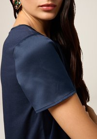 Top corto blu navy a maniche corte con texture liscia; scollatura tonda e morbido drappeggio. Accento notevole dato da orecchini dorati con gemme colorate.