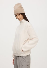 Giacca in pile di colore crema chiaro con colletto alto, zip completa e tasche laterali. Presenta un cordino elastico all'orlo.