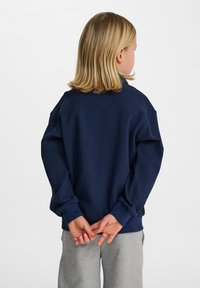 Marinblå texturerad sweatshirt med avslappnad passform, ribbade muddarn och rundad nederkant, som bärs över ljusgrå byxor. Bakifrån med händerna knäppta.