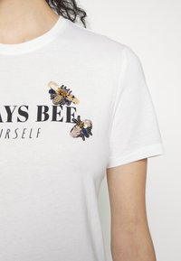 Valkoinen puuvillapaita, jossa on beigellä, kultaisella ja mustalla brodeeratut mehiläiset sekä mustalla tekstillä painettu "ALWAYS BEE" -viesti. Lyhyet hihat.