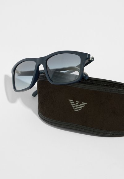 Μπλε πλαστικά γυαλιά ηλίου με gradient φακούς, resting πάνω σε ένα καφέ φουτζαιο suede θήκη με ασημένιο λογότυπο. Λεπτός σχεδιασμός και στρογγυλεμένες άκρες.