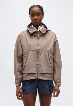 Nő bézs kapucnis dzsekiben, amely fel van húzva egy fehér felső és sötétkék sportnadrág fölé, egyszerű háttér előtt áll.