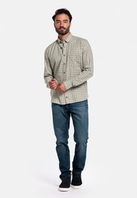 Lichtgroene geblokte overhemd met knoopsluiting en lange mouwen, gecombineerd met blauwe spijkerbroek en donkere sneakers. Eenvoudige achtergrond vergroot de zichtbaarheid.