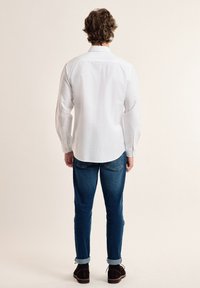 Camisa blanca de manga larga con cuello abotonado, detalle de yugo en la parte posterior, combinada con jeans de mezclilla azules y zapatos de ante marrones.