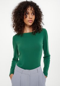 Zielony bodysuit z długim rękawem, wykonany z gładkiego materiału, z okrągłym dekoltem, w zestawie z jasnoszarymi spodniami o wysokim stanie, zdobionymi plisami.