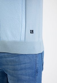 Suéter azul claro con diseño de tejido texturizado, dobladillo acanalado y logotipo bordado en negro en el costado, combinado con pantalones de mezclilla azules.