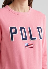 Rosa sweatshirt med ribbad rundhals, med broderade bokstäver "POLD" i marinblått och en amerikansk flagga som patch nedanför. Mjuk textur.