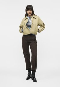 Γυναίκα που φοράει μπεζ cropped μπουφάν, μπλε και καφέ ριγέ τοπ, σκούρο καφέ παντελόνι και μαύρα μποτάκια με τακούνι, στέκεται με τα χέρια στις τσέπες.