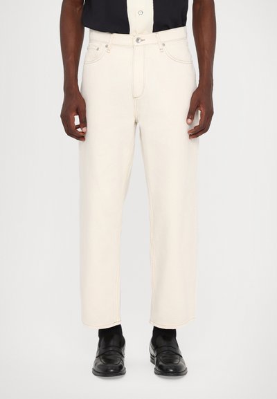 Pantalons en denim couleur crème avec une coupe droite, présentant des coutures visibles et des poches avant. Portés avec des chaussures noires.