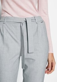 Grijze op maat gemaakte broek met een gestructureerde afwerking, voorzien van een brede riem en zijzakken. Gecombineerd met een lichtroze top met lange mouwen.