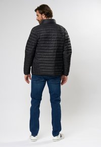 Schwarze Pufferjacke mit hohem Kragen, gesteppter Textur und taillierter Silhouette. Kombiniert mit blauen Jeans und weißen Sneakers.