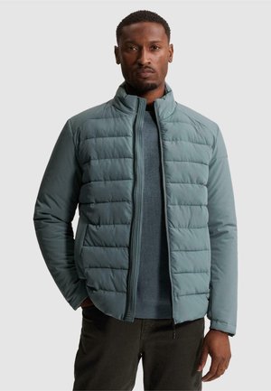 Homme portant une veste matelassée bleu clair sur un pull bleu avec un pantalon noir, debout avec une main dans la poche devant un fond uni.