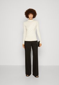Hvid langærmet top med draperet design, parret med høj talje sorte vide bukser. Holder en lille sort clutch.