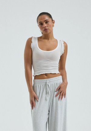 Femeie cu părul prins, purtând un top alb fără mâneci, cu volane și pantaloni sport gri deschis cu șnur, stând pe un fundal simplu.