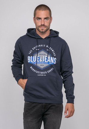 Homme aux cheveux courts portant un sweat à capuche bleu marine avec le texte « BLUE & JEANS » et un jean noir, debout devant un fond clair uni.