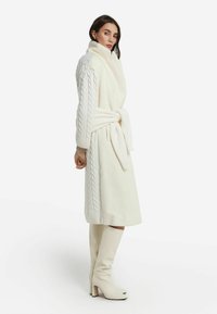 Cappotto bianco avvolgente con maniche a treccia e vita regolabile con una cintura. Ha un collo alto ed è abbinato a stivaletti con tacco al ginocchio.