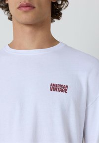 Bijela pamučna majica s natpisom "AMERICAN VINTAGE" u tamnocrvenoj boji na lijevoj strani prsiju. Klasičan okrugli izrez, mekani materijal i opušteni kroj.