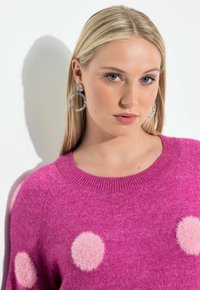 Blonde Frau mit blauen Augen, die marmorierte Creolen und einen knallpinken Pullover mit hellrosa flauschigen Punkten trägt, vor weißem Hintergrund.