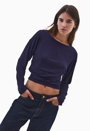Haut violet à manches longues avec un décolleté large, un corsage ajusté et un tissu texturé, associé à un jean en denim foncé avec des garnitures dorées.