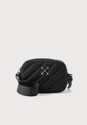 Bolso negro acolchado cruzado con un emblema plateado, cierre de cremallera y una correa ancha con la marca, sobre un fondo liso.