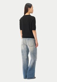 Kvinna stående i profil, klädd i en svart kortärmad tröja och högmidjade blekta blå vida jeans med synliga bakfickor.