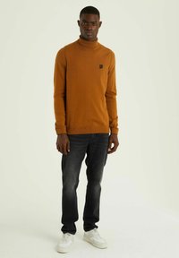 Maglione a collo alto marrone con un fit slim, realizzato in un materiale morbido, abbinato a jeans scuri e sneakers bianche. Presenta un piccolo logo applicato.