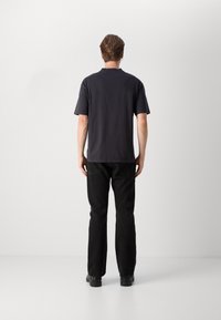 Homme debout, dos tourné, portant un t-shirt noir à manches courtes, un pantalon noir et des chaussures noires sur un fond blanc uni.