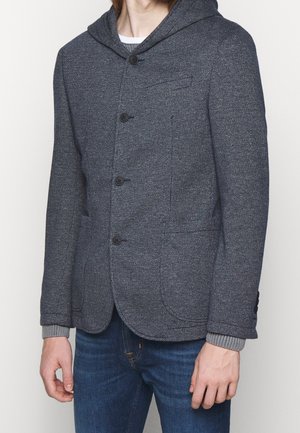 Homme portant un blazer à capuche texturé gris foncé avec quatre boutons, des poignets de pull gris clair et un jean bleu sur fond uni.