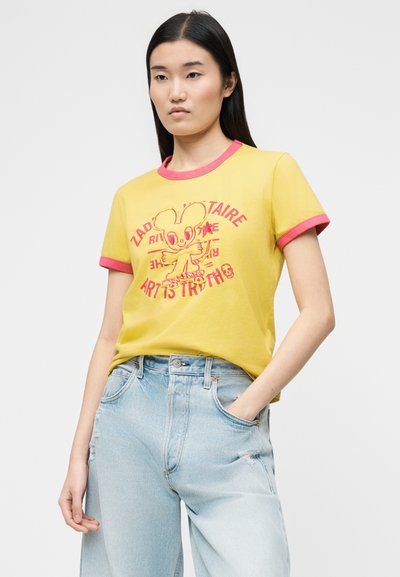 Νεαρή γυναίκα που φοράει κίτρινο μπλουζάκι με ροζ περιθώριο και γραφικό καρτούν ποντικιού, συνδυασμένο με ανοιχτό μπλε τζιν με σκισίματα, όρθια με το ένα χέρι στην τσέπη.