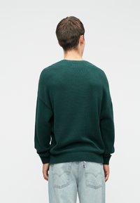 Zeleni pleteni pulover z ohlapnim krojem, rebrastimi manšetami in robom. Ima okroglo ovratnico in teksturirano tkanino, v kombinaciji s svetlimi kavbojkami.