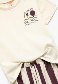 Cremefarbenes Baumwoll-T-Shirt mit einem Surfer-Graphic, kombiniert mit braun-beigen gestreiften Shorts mit elastischem Bund und Kordelzug.