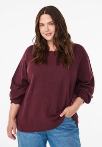 Burgunderfarbener Oversized-Pullover mit fallenden Schultern und gerippten Bündchen, kombiniert mit hellblauen Jeans. Weicher, strukturierter Stoff mit lässiger Passform.