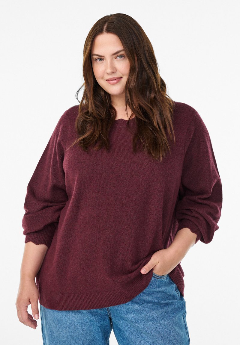 Burgunderfarbener Oversized-Pullover mit fallenden Schultern und gerippten Bündchen, kombiniert mit hellblauen Jeans. Weicher, strukturierter Stoff mit lässiger Passform.