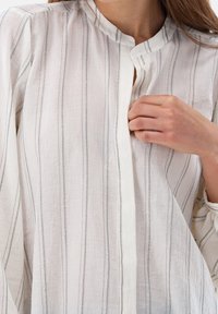 Chemise en lin blanc avec des rayures grises verticales, dotée d'un col montant et d'une fermeture à boutons, mettant en valeur un tissu doux et texturé.