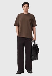 Bruine katoenen t-shirt met een relaxte pasvorm, korte mouwen en een klein logo op de borst, gecombineerd met losse zwarte broeken en een zwarte tas.