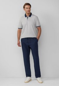 Herren Poloshirt mit kurzen Ärmeln in Weiß mit Rauten-Muster, dunkelblauem Kragen und Ärmelsäumen; kombiniert mit dunkelblauen Jogginghosen mit Kordelzug und weißen Sneakers.