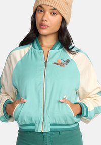 Teal bomber jacket with cream sleeves, ribbed cuffs, and a decorative embroidered design featuring a sun and bird. Zippered front and pockets.