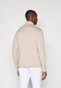 Beež pikkade varrukatega särk kõrge kaelusega, sileda tekstuuriga ja tagasi õrn logo detailiga, kantud valgete pükstega.