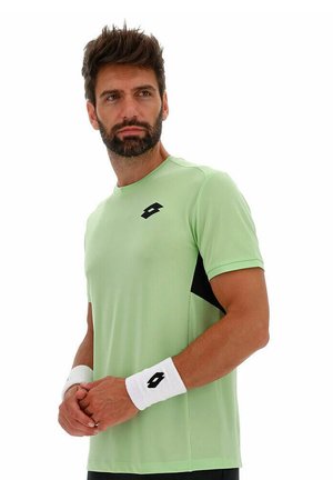 Mann mit Bart, der ein hellgrünes Sportshirt und weiße Handgelenkbänder mit schwarzen Logos trägt, steht mit verschränkten Händen da und schaut zur Seite.