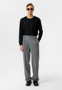 Zwarte langemouwen shirt gecombineerd met hooggesneden, geplooide grijze broek. Model draagt zwarte ronde zonnebril en stevige zwarte schoenen.