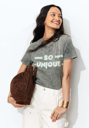 Femme souriante aux longs cheveux foncés portant un t-shirt gris "SO UNIQUE" et un pantalon blanc, tenant un sac marron à imprimé léopard sur le bras.