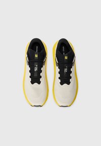 Baskets de sport beige clair avec une semelle jaune et des accents noirs. Présente un dessus texturé, des lacets et une étiquette sur la languette.